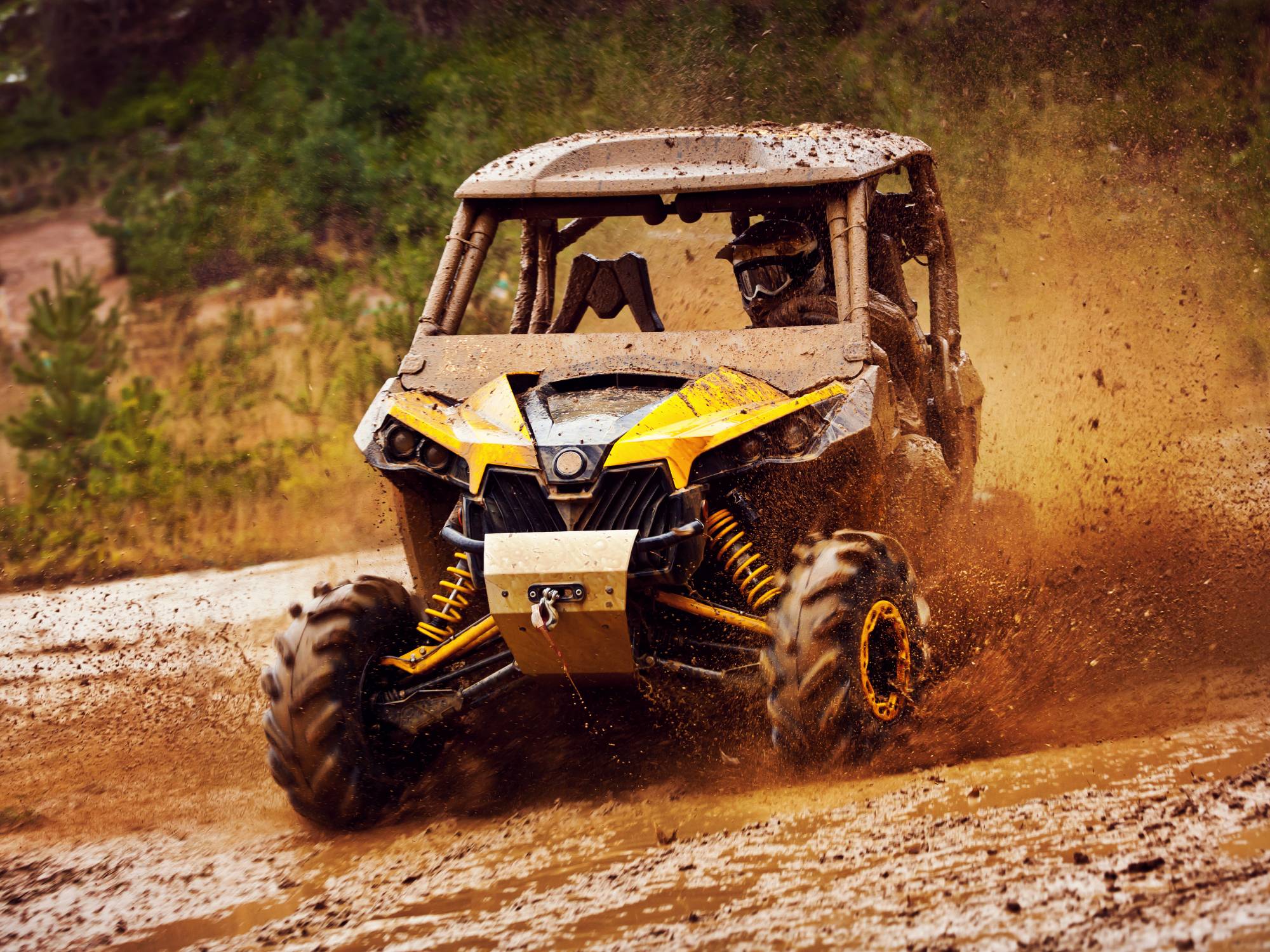 A driver wearing a helmet and goggles, riding in a yellow UTV, and kicking up mud and dirt on a rugged off-roading trail.