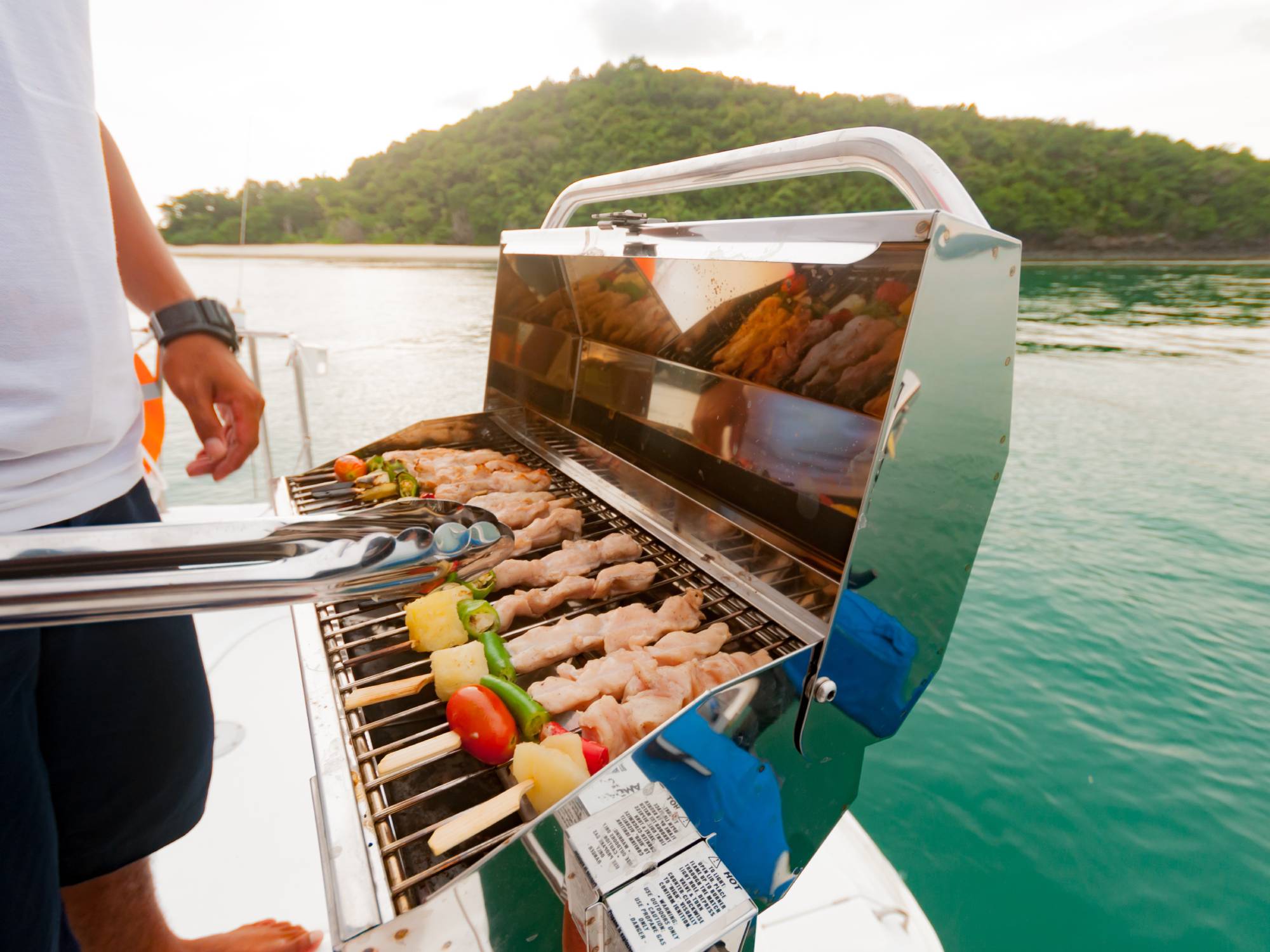 A sleek chrome grill is cooking up some delicious looking skewers. The grill is on a boat in the middle of a gorgeous lake.