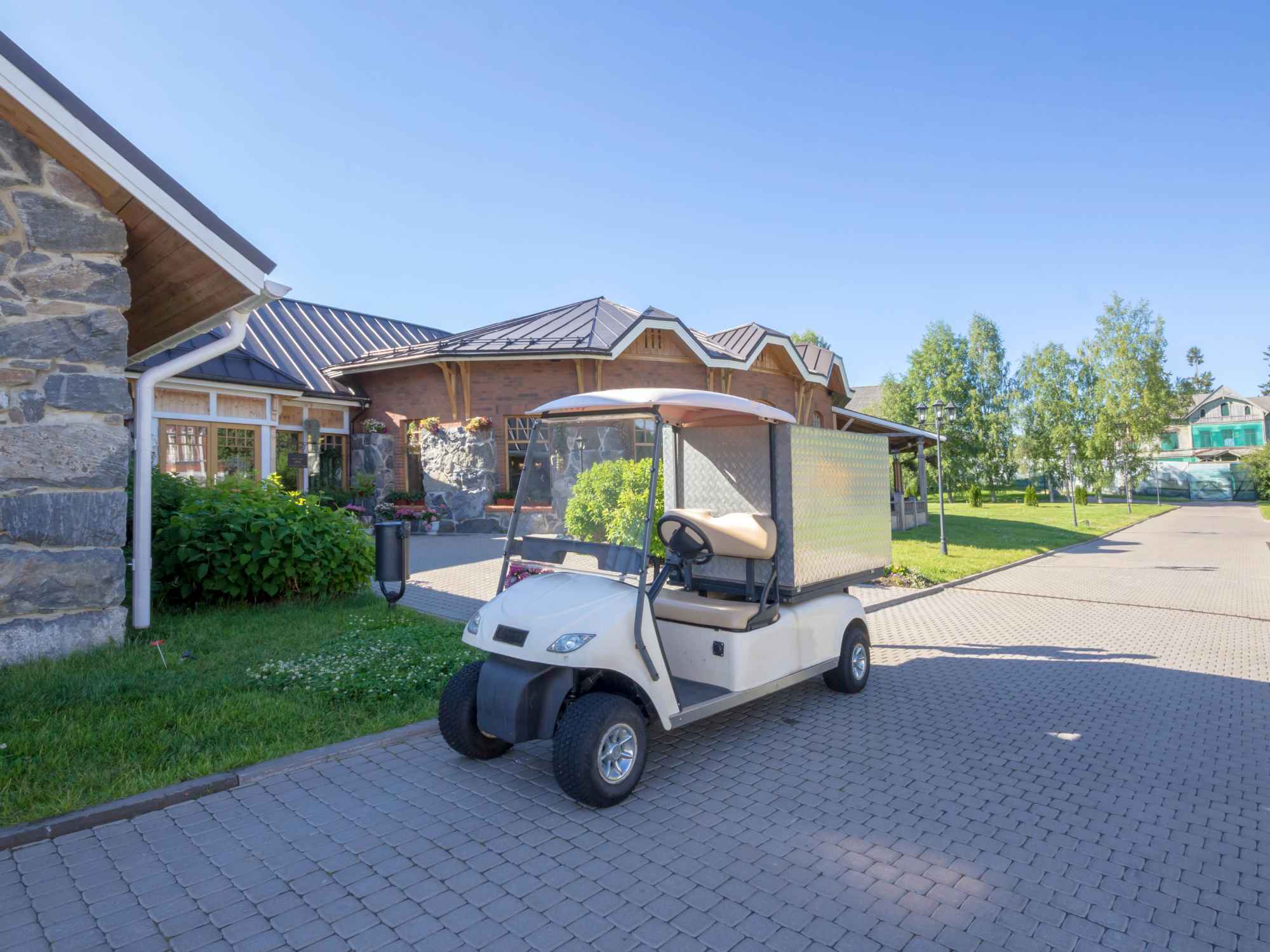 A white golf cart is parked in front of a house in a residential area. The cart is in the shade to protect it from the sun.
