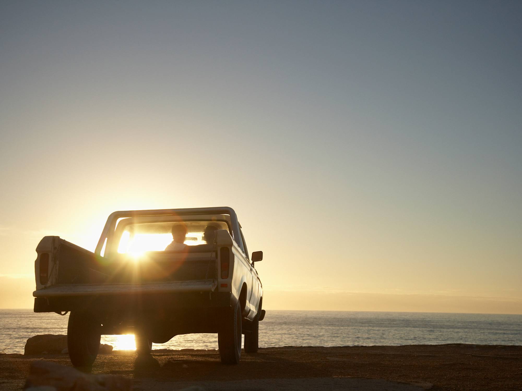 A pick-up truck facing a beautiful sunset over a large body of water. A couple sits inside the cabin, enjoying the view.