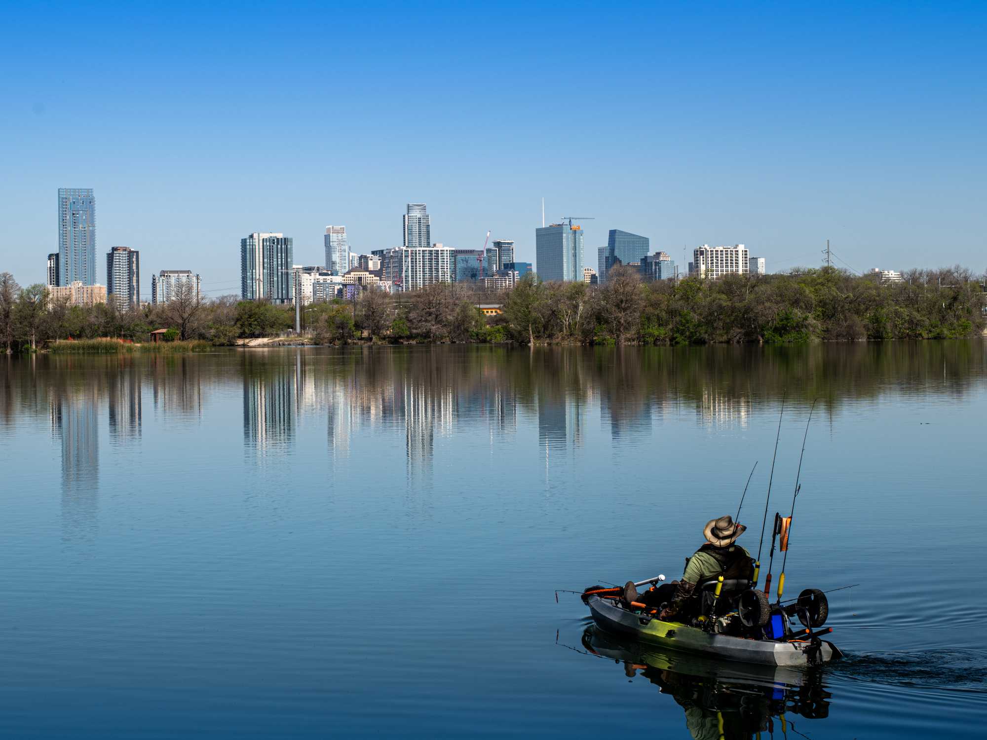 An angler sitting with poles and fishing equipment in a kayak on a lake with the Austin, Texas, skyline in the background.