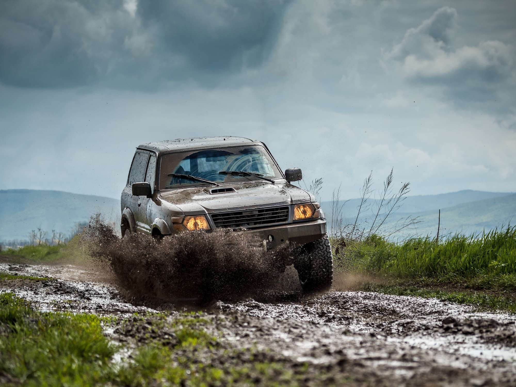 A sport utility vehicle driving through the mud while off-roading on a cloudy day. There are mountains in the background.