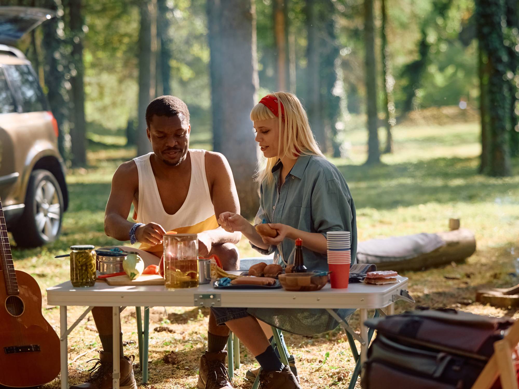 A man and a woman sitting at an outdoor picnic table in a wooded area. They are preparing a meal together.