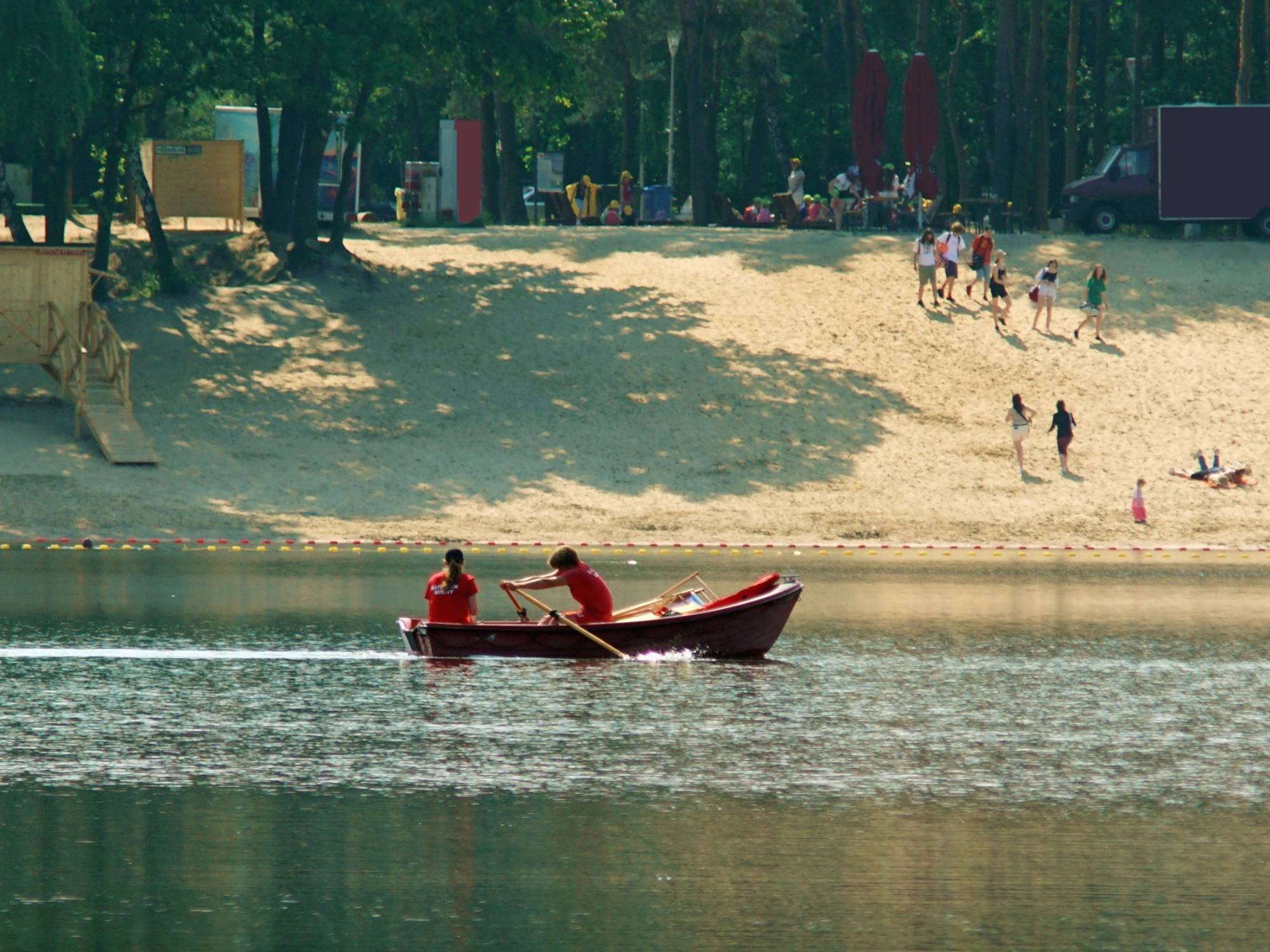 Two lifeguards in red shirts sit in a canoe while people walk on the beach at the edge of a lake.