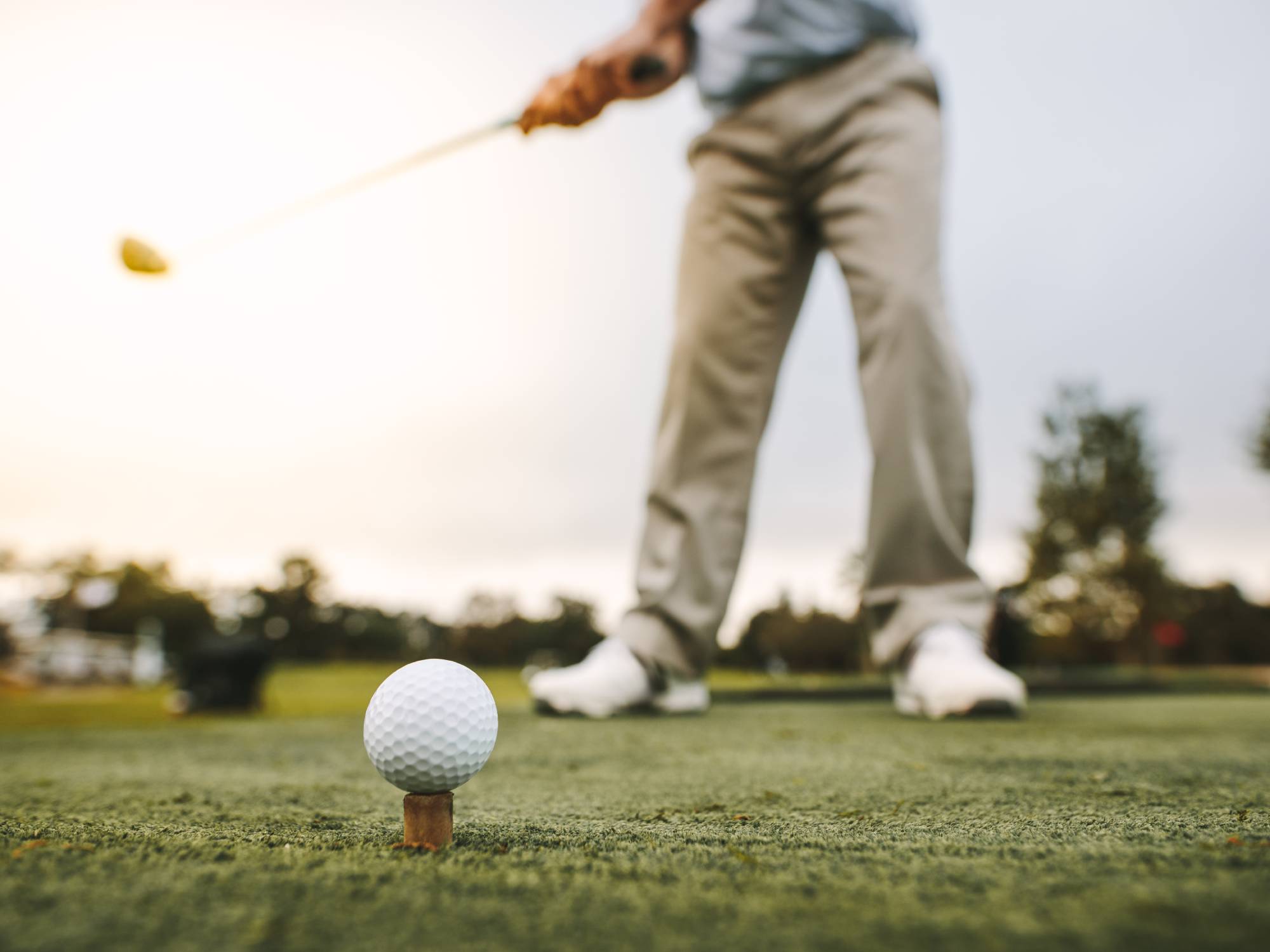 A golf ball and a tee on a green with a man standing behind it getting ready to hit it with a golf club.