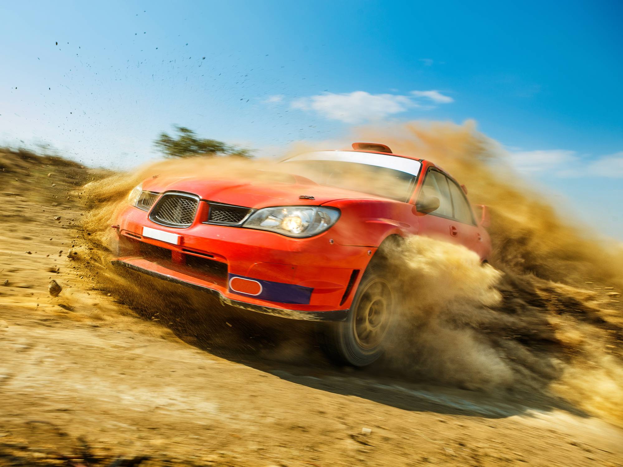 A front view of a red rally race car drifting through a dirt track while kicking up dirt all over the car and track.