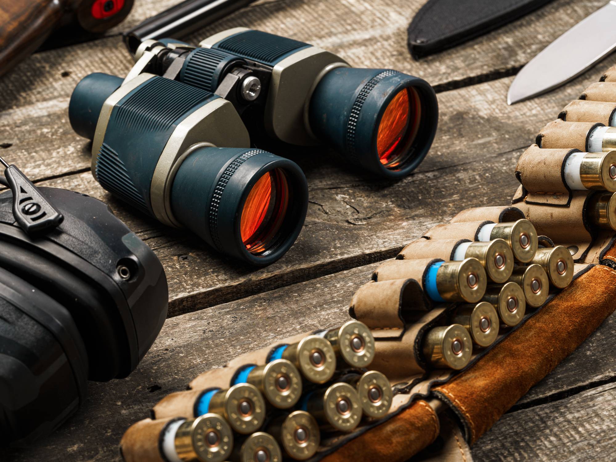 Assorted hunting equipment, including binoculars, bullets in a leather case, and knives, on a wooden surface.