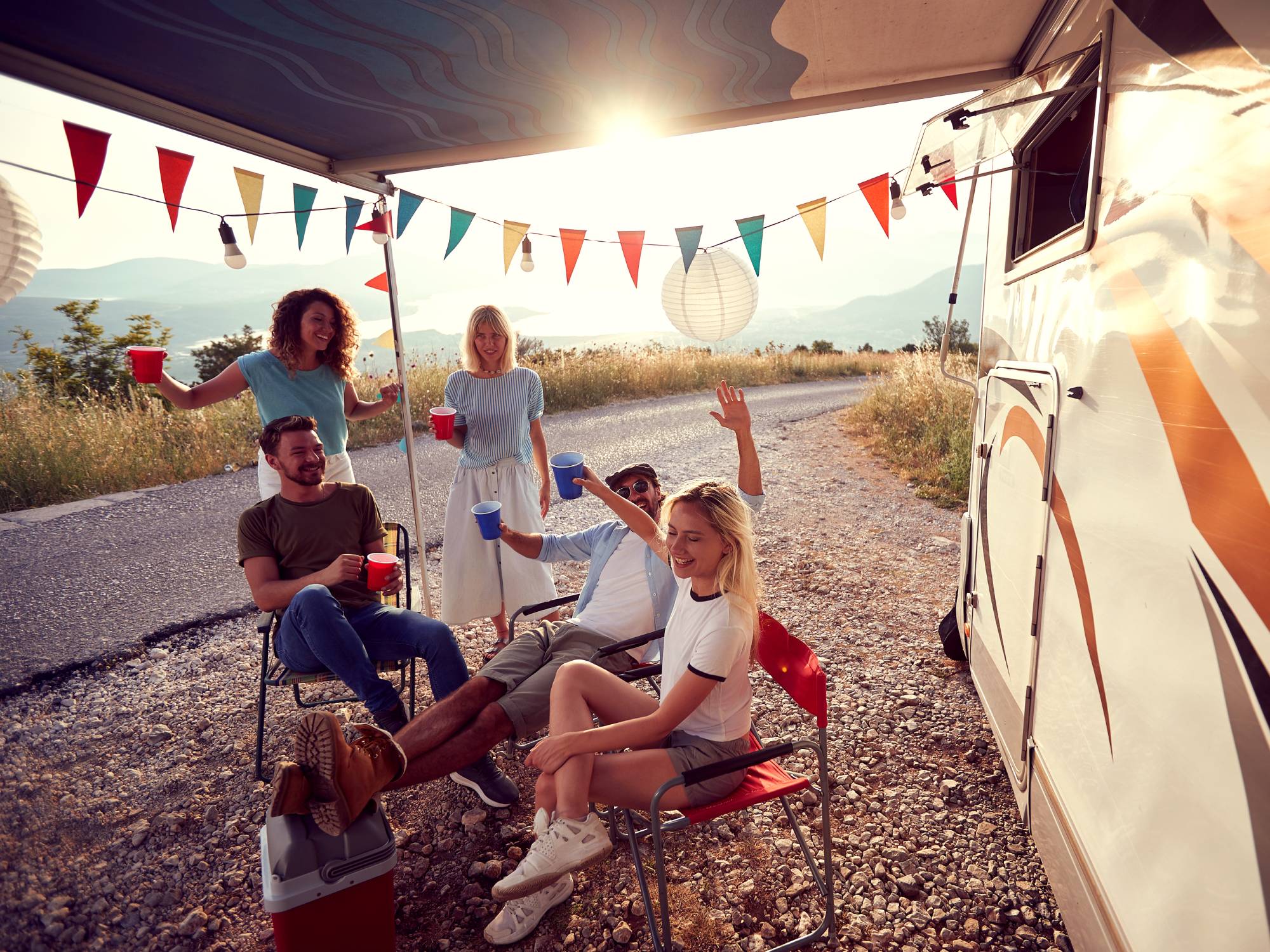 A group of friends sitting outside of an RV with the overhang open and banner pennants hanging from the side.