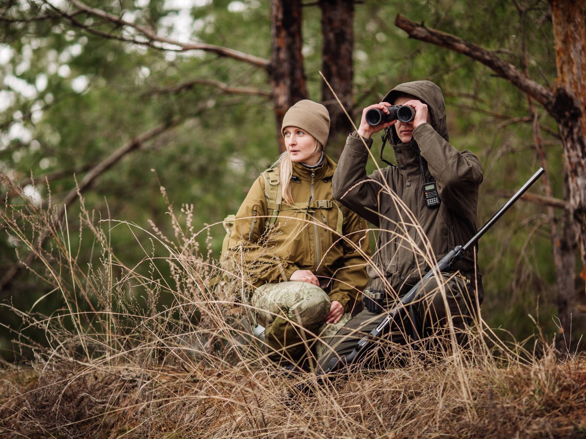 A man and woman are crouched down in a forest. They hold binoculars and a hunting rifle. Their clothing helps them camouflage.