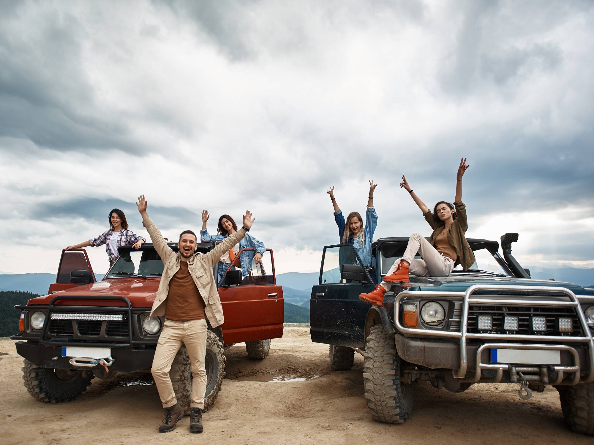 Five people smiling with their arms in the air. The women stand on two off-roading trucks and a man stands on the dirt.