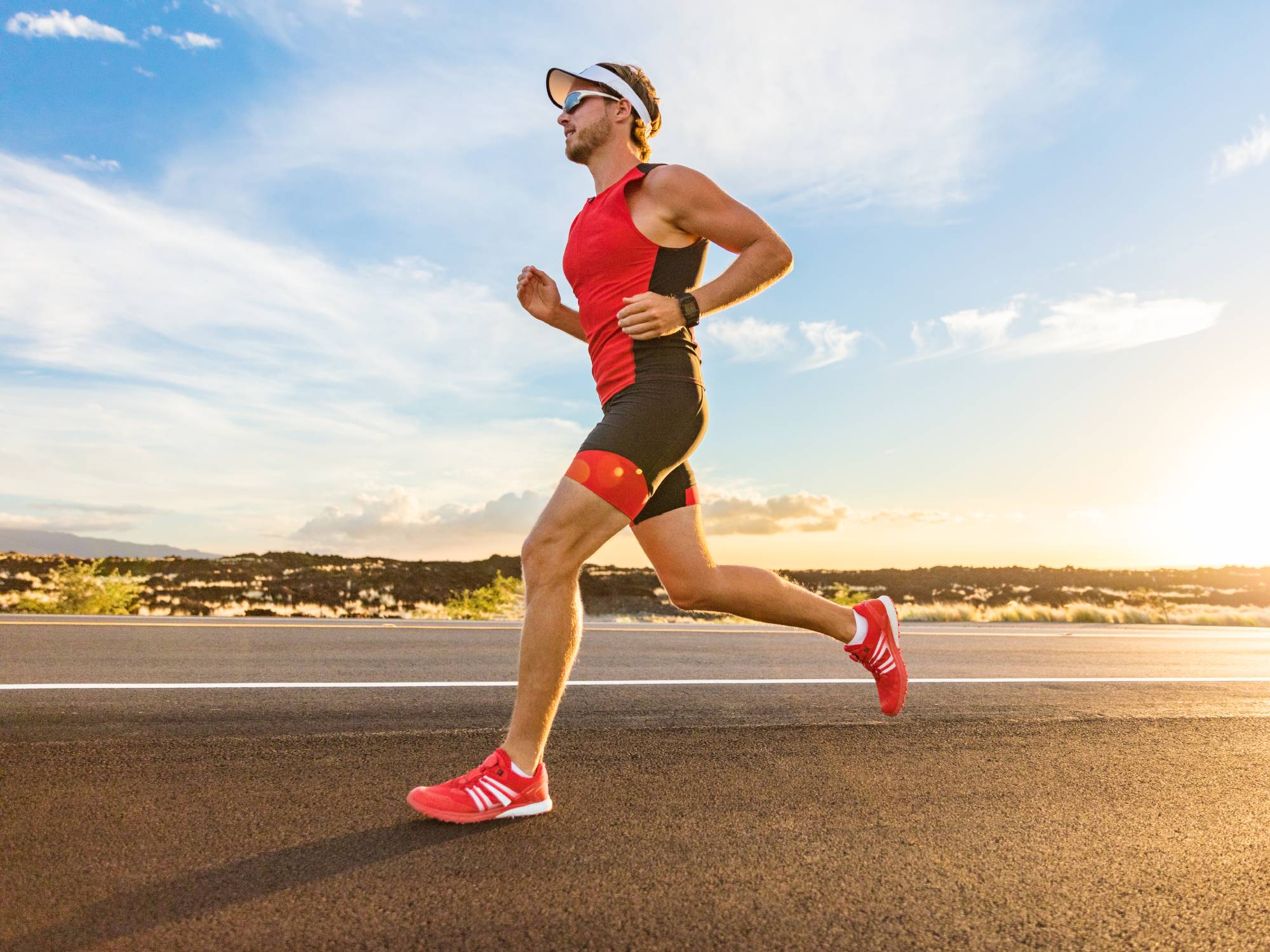 A person wearing a red and black workout set, red sneakers, a hat, and sunglasses runs outside.