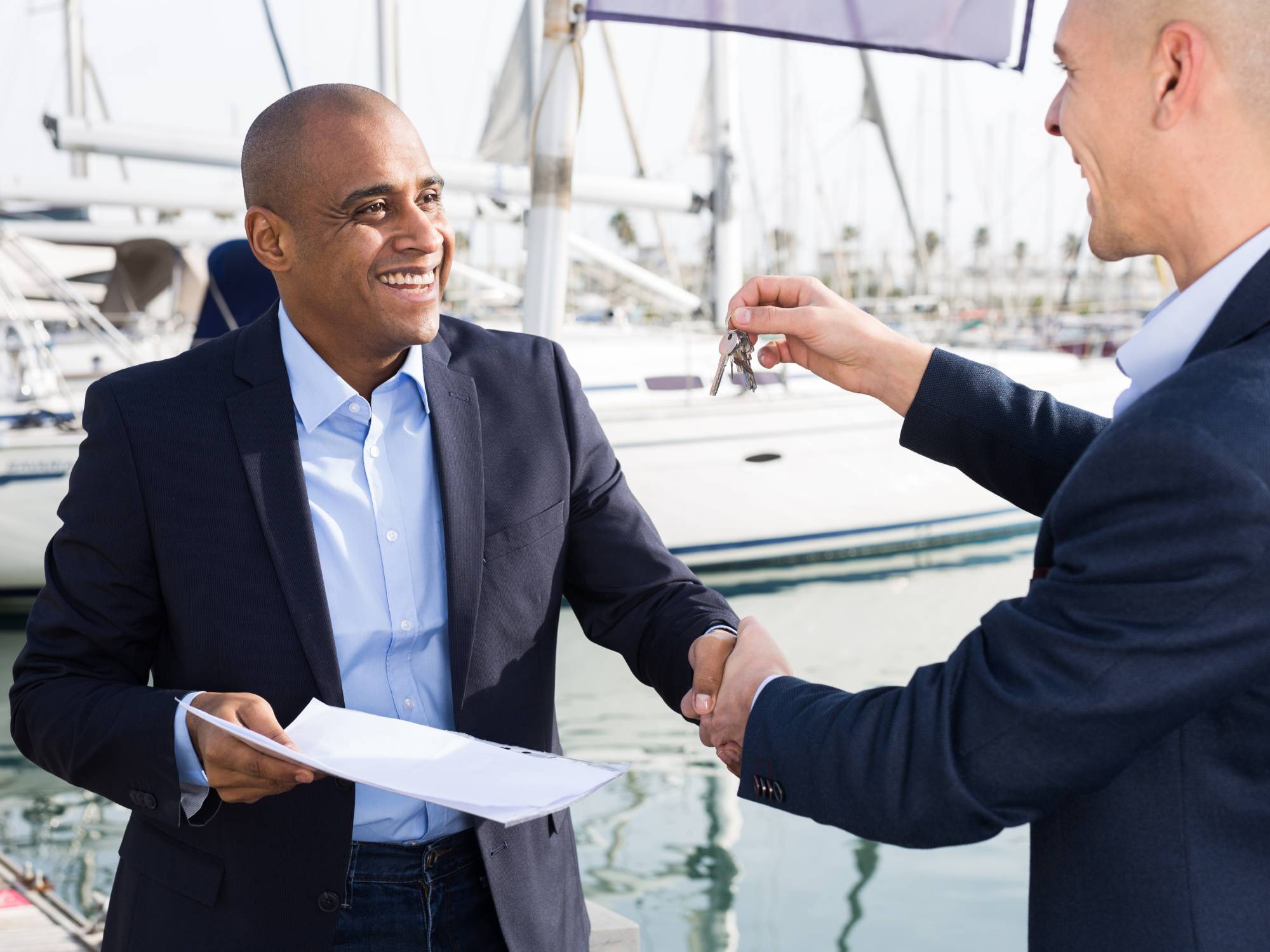 Two men in professional clothing shaking hands on a marina dock with boats in the water. One man holds out a key.