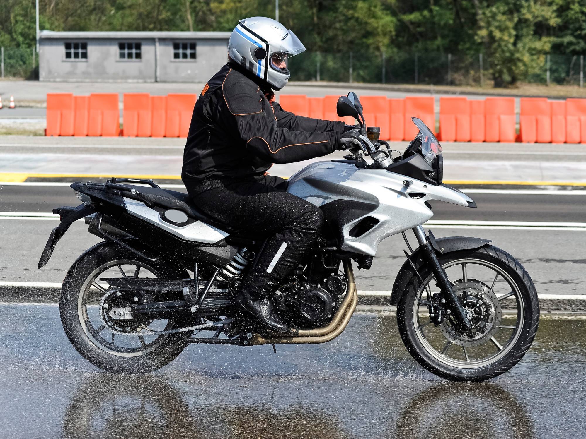 A man wearing a silver helmet and a black outfit rides a silver motorcycle down a road on a rainy day.