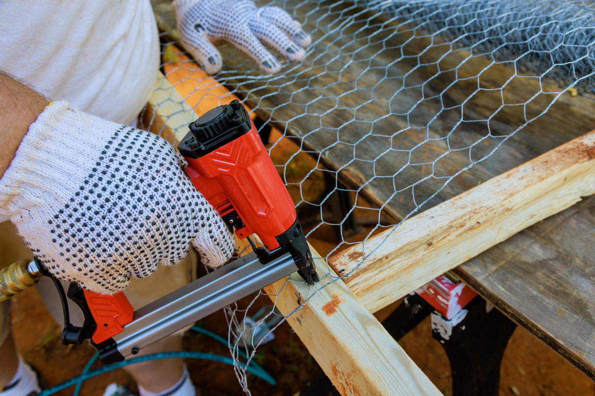 A worker in white gloves uses a red staple gun to attach chicken wire to a wooden frame on a workbench.