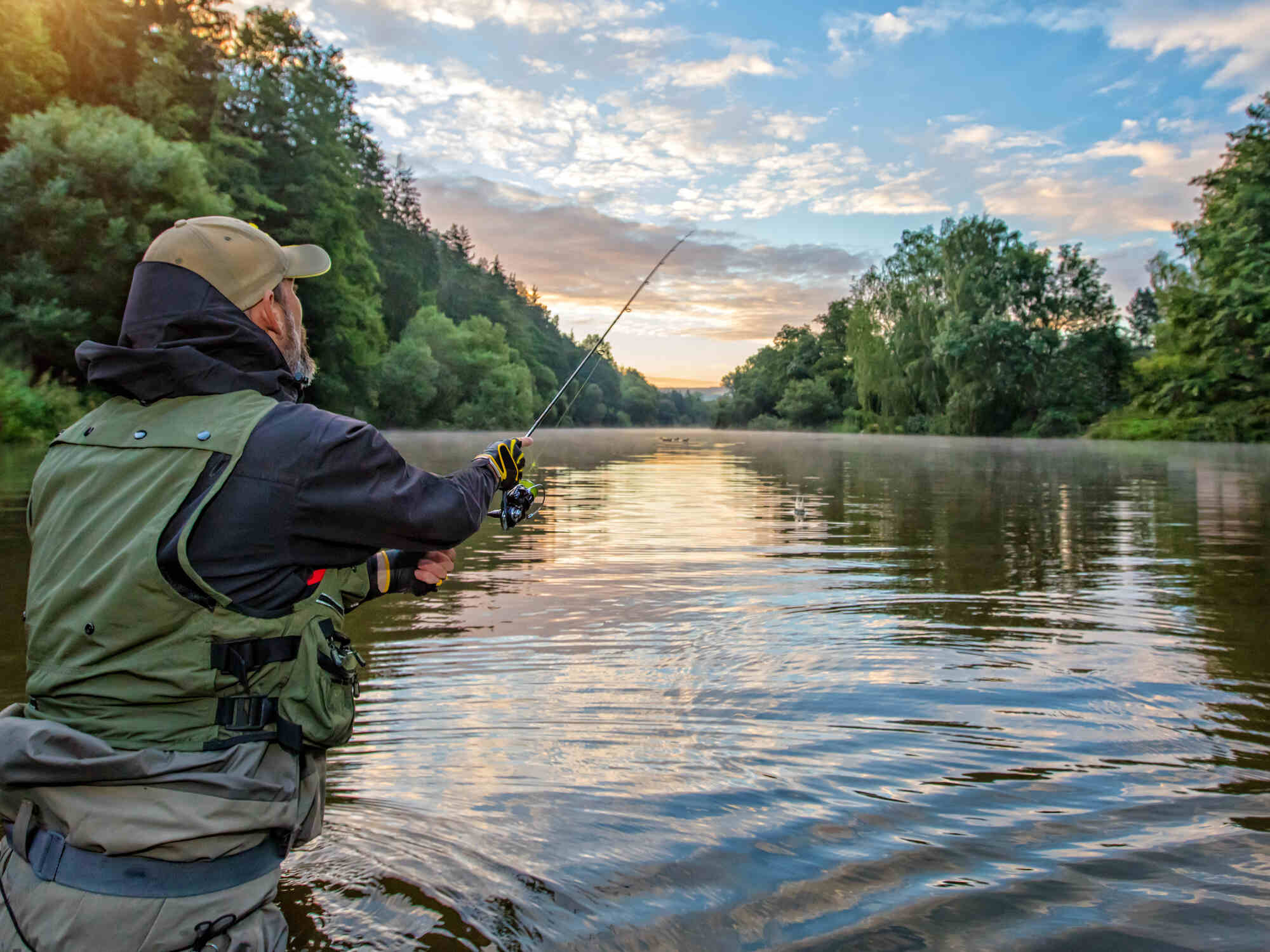 Rear view of a man wearing waders and standing in a river while fishing. Trees and a clear sky are in the background.