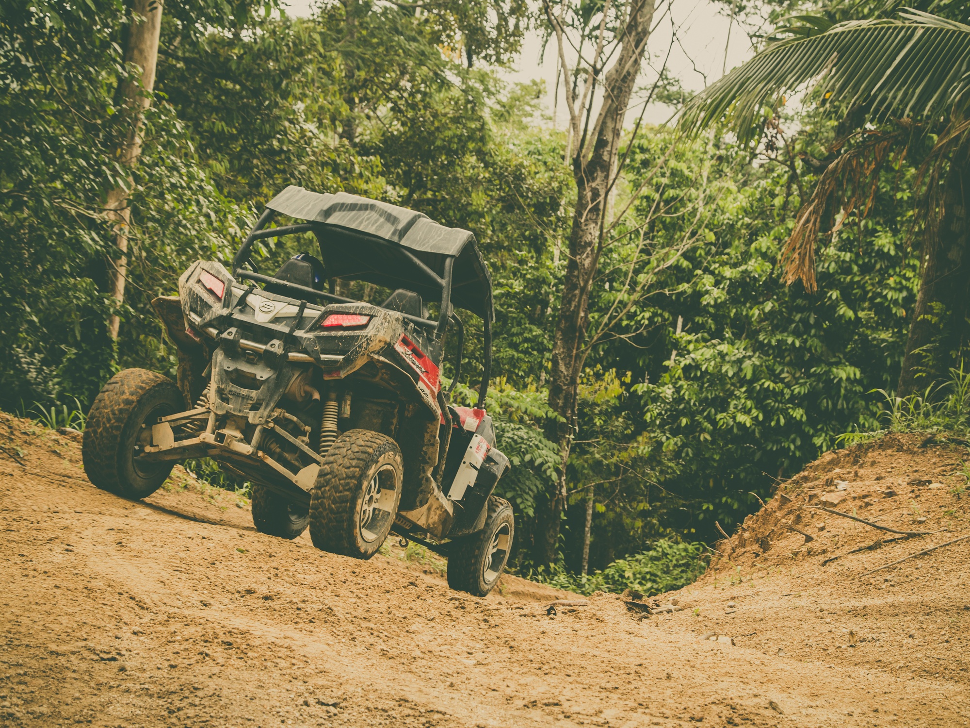 A large off-road vehicle with a raised body stands on an inclined dirt road in the middle of a forest.