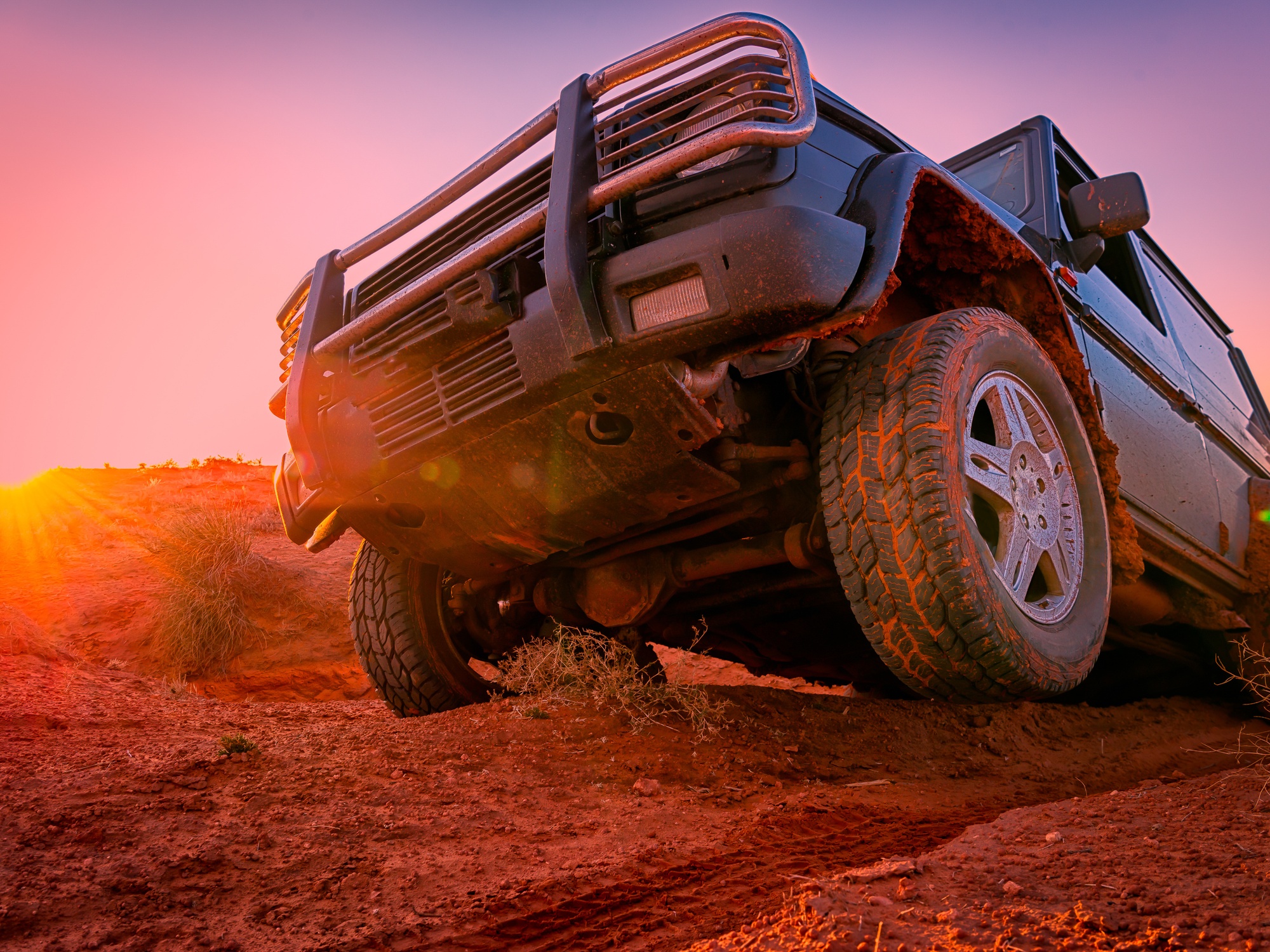 A close up of an off-road vehicle crawling through the desert at sunset. The sun is bright orange behind the vehicle.