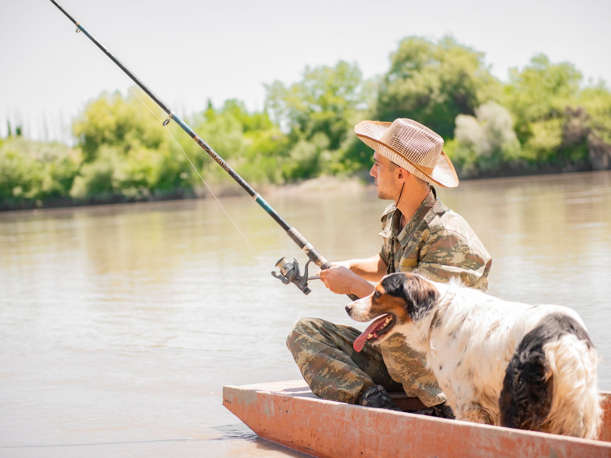 A man and his dog are in a canoe, fishing in a brown lake edged by trees on a hot, sunny day.