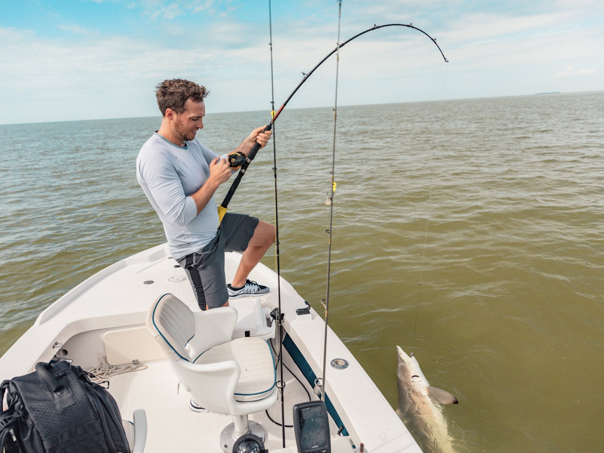 A man stands on a smaller boat in the middle of a body of water. He is reeling in a fish with his rod.