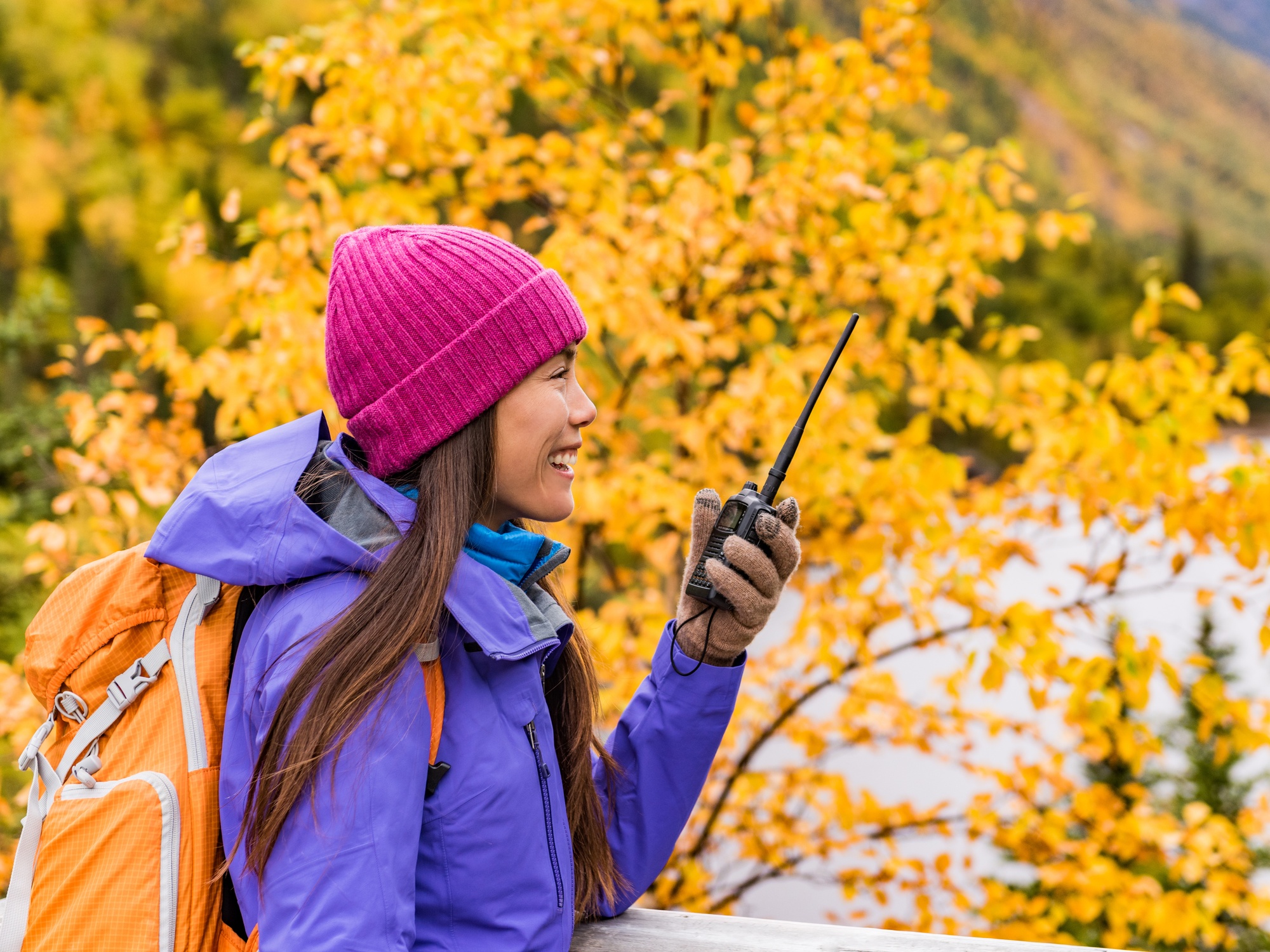 A woman in a purple jacket and pink beanie carries an orange backpack and a radio while standing outside.