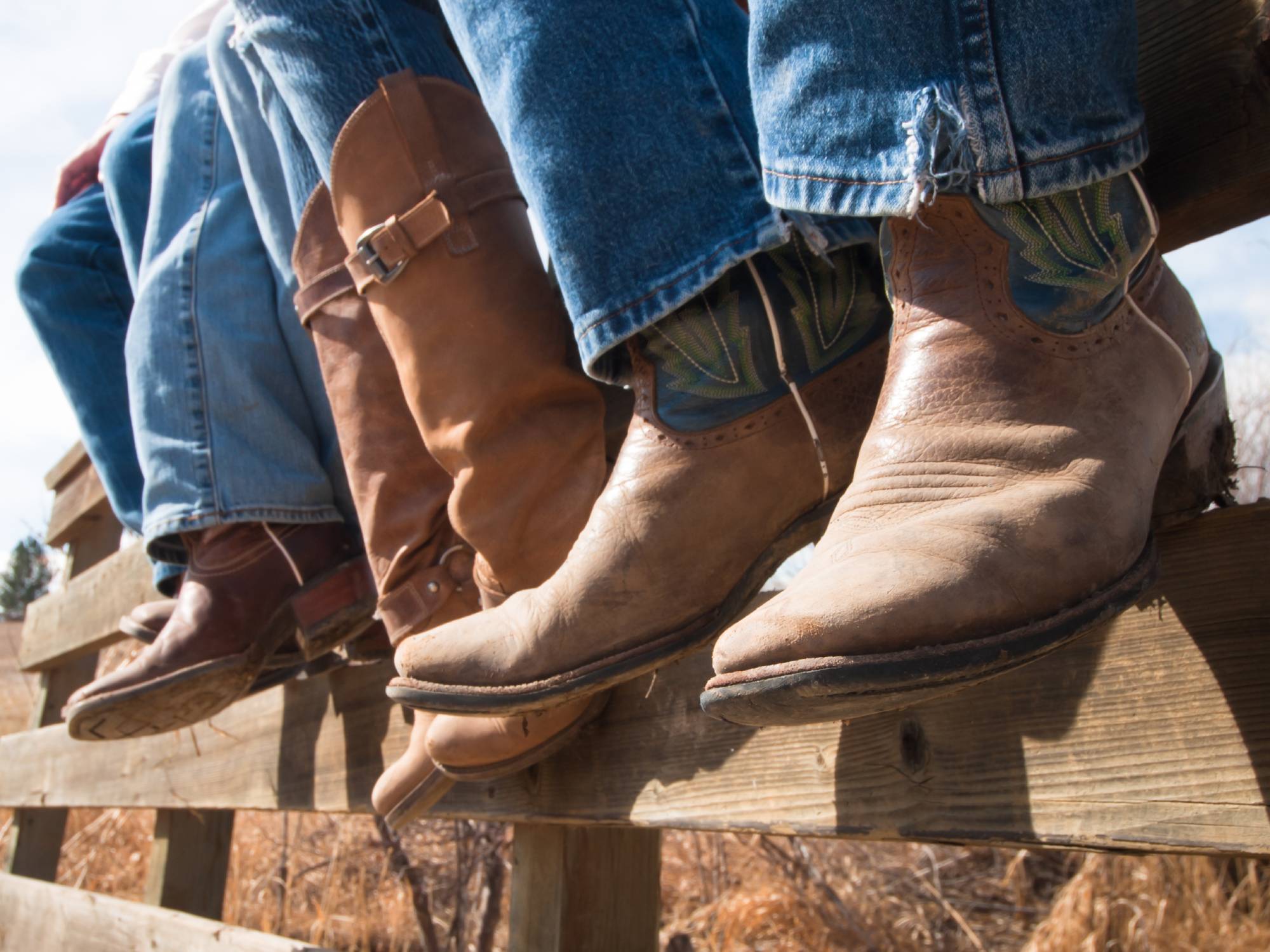 The bottom of three people's jeans and their cowboy boots, while they sit outside on top of a wooden fence.