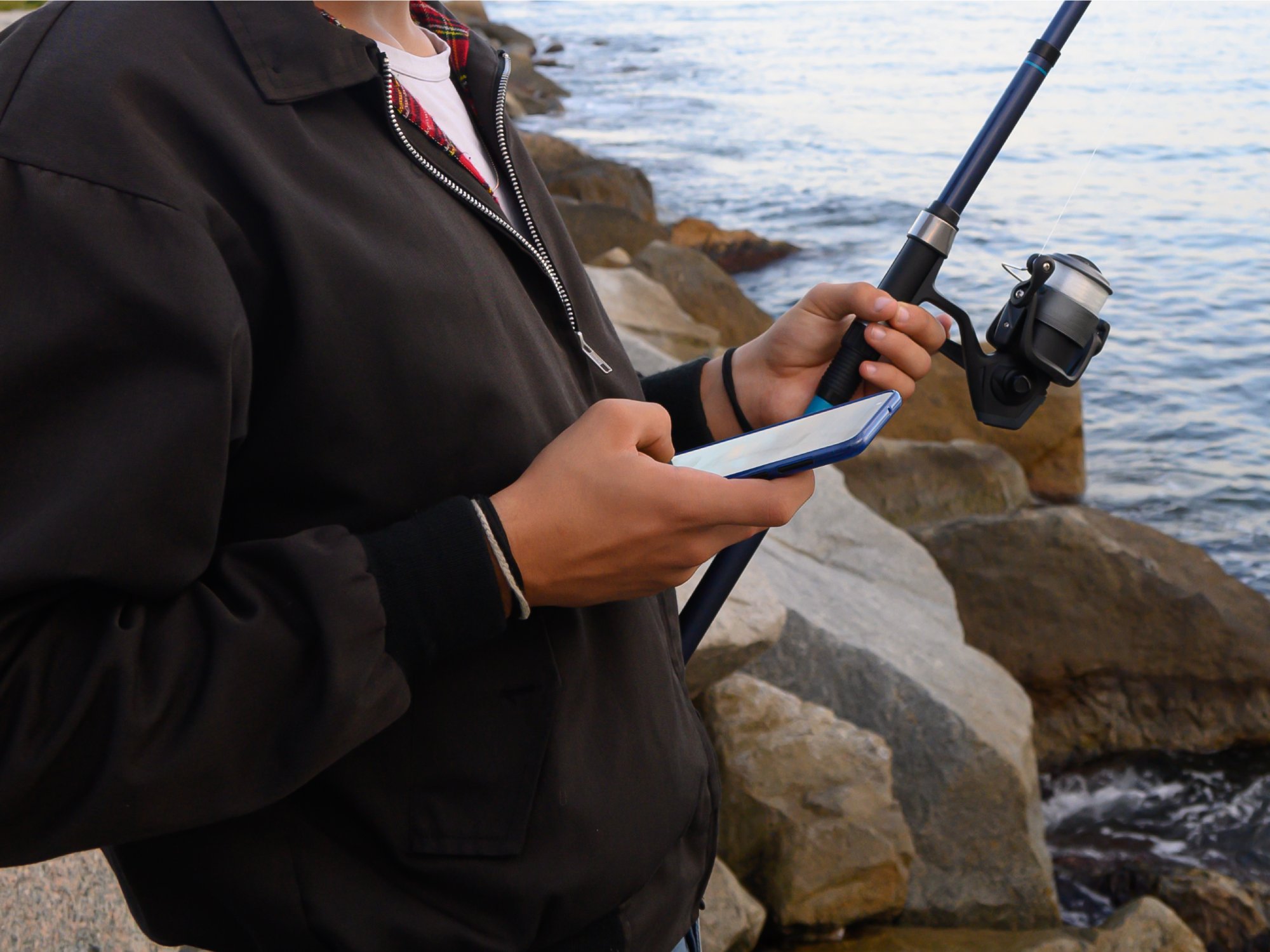 A close-up view shows a person holding a phone in one hand and a fishing pole in the other while standing on a rocky shore.