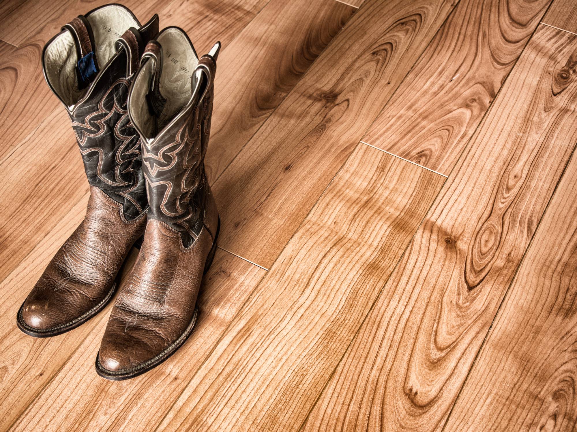 A pair of old cowboy boots made of worn leather, with visible creases and scuffs, sitting side by side on a hardwood floor.