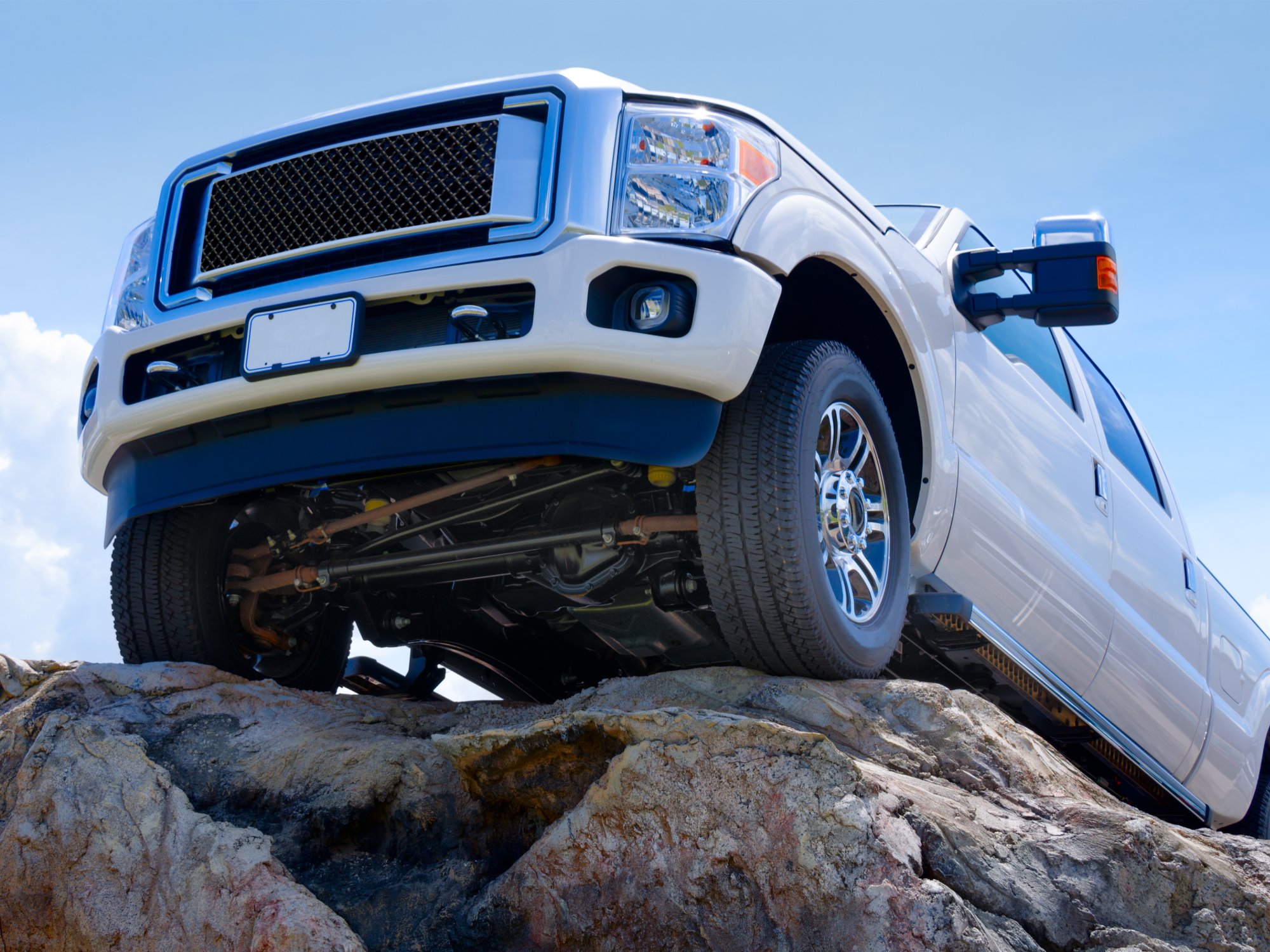 A white truck sits on the edge of a rock. The sky is blue in the background behind the vehicle, with white clouds.