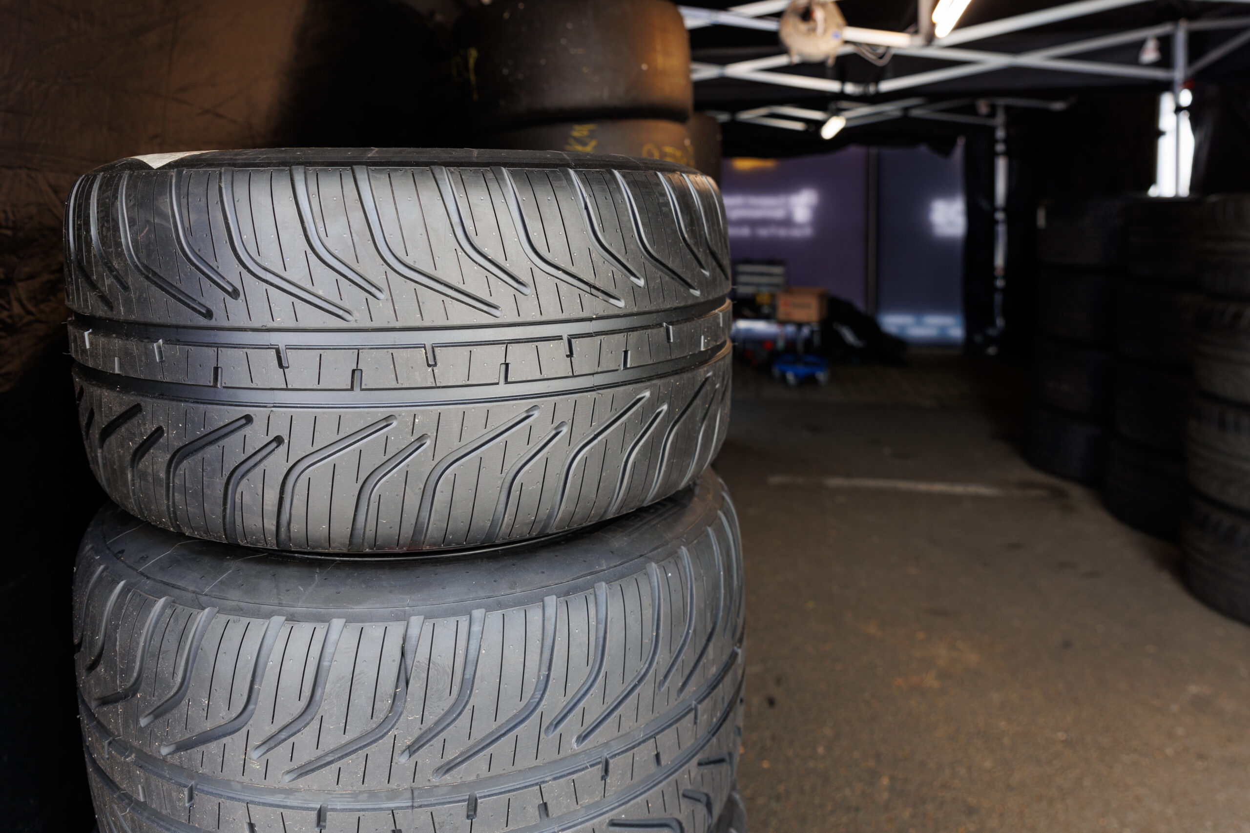 Close-up of wide black tires stacked in an auto shop, showing patterned tread and rows of tires behind them.