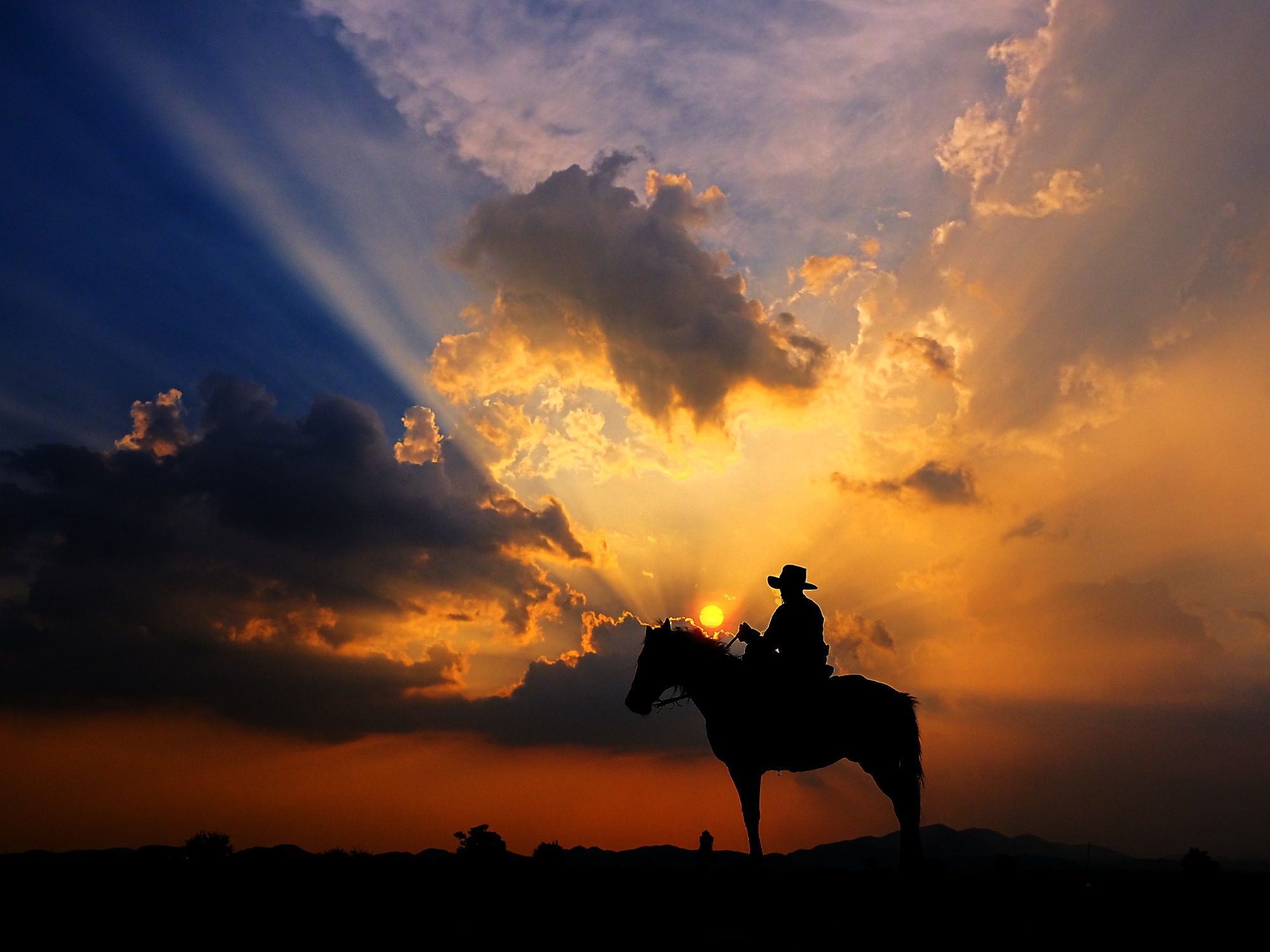 A silhouetted hat-wearing rider on a horse against an orange sunset sky with clouds and radiating sun rays.
